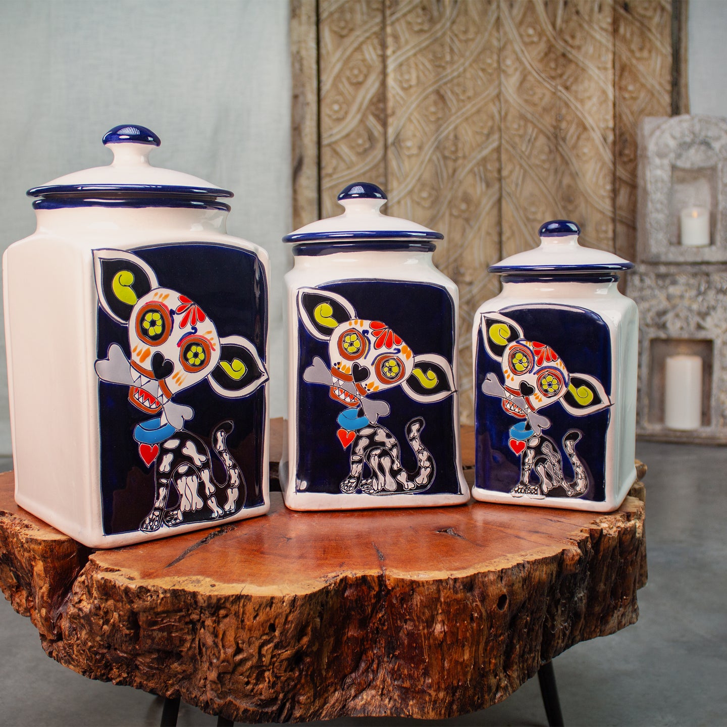 Three white ceramic canisters with blue lids. Hand painted with colorful day of the dead dog illustrations on a dark navy background. On a rustic wood coffee table.