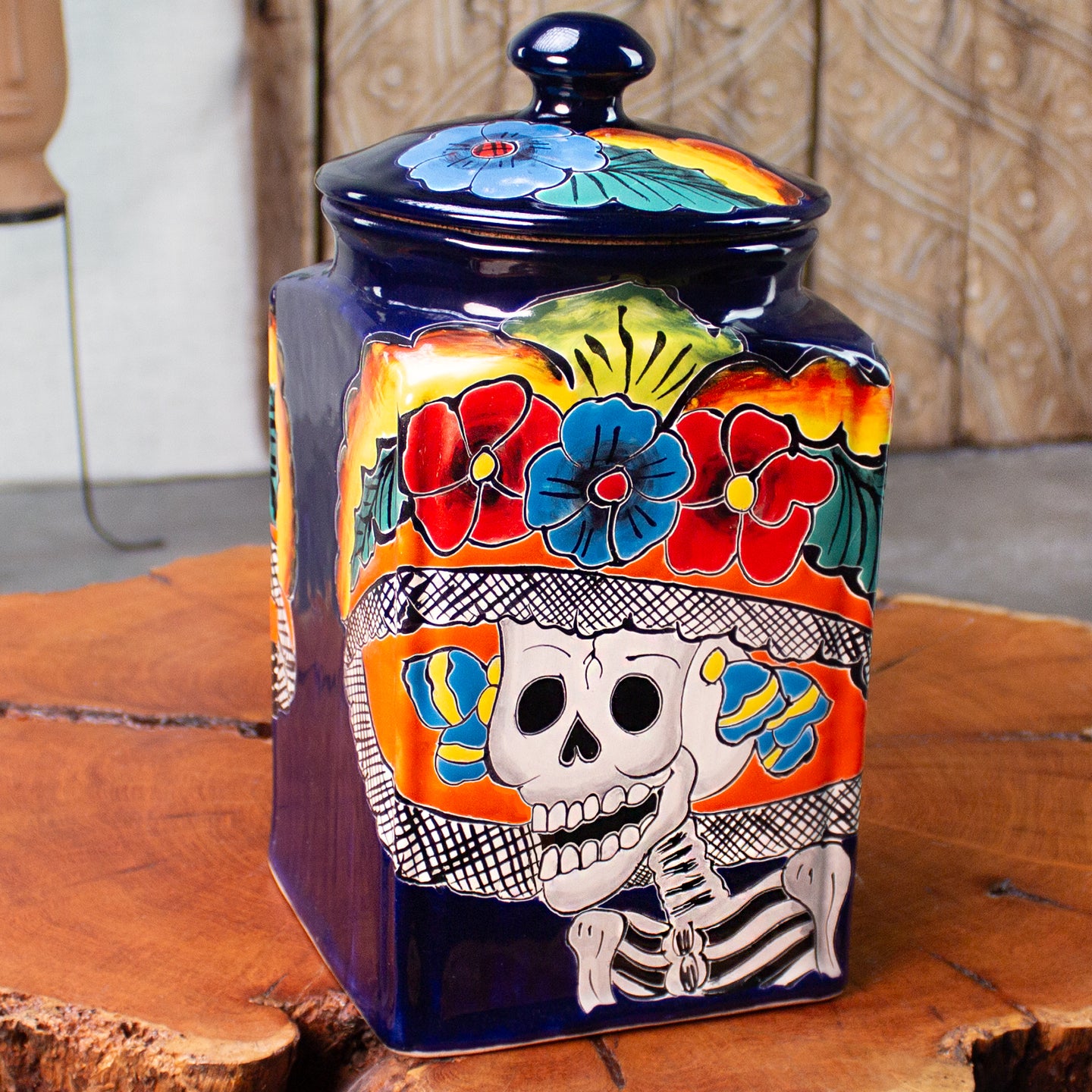 A square ceramic jar with a matching lid, decorated with a colorful Day of the Dead style design featuring a white skeleton, red and blue flowers, and a blue background, displayed on a rustic wood table.