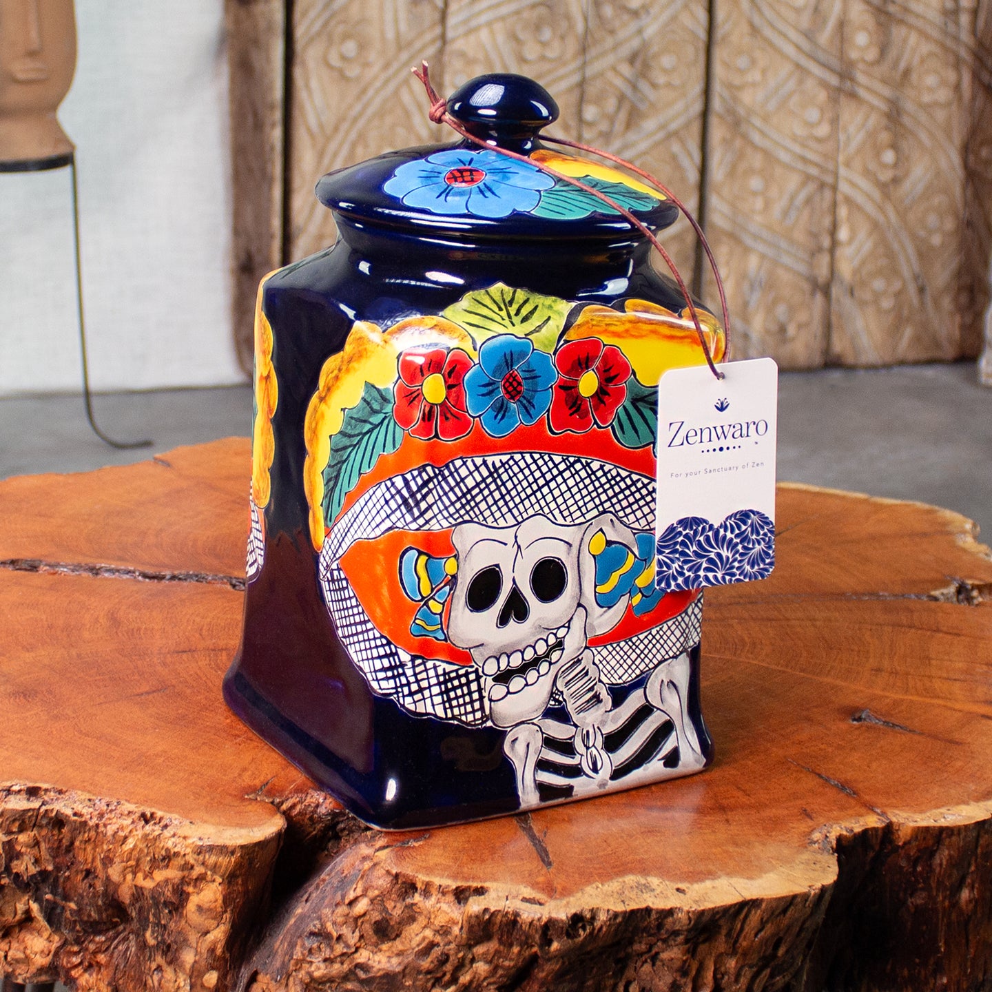 A hand-painted ceramic Day of the Dead (Día de los Muertos) jar with a lid, featuring a sugar skull design and colorful floral motifs, on a rustic wood table. A 'Zenwaro' hang tag is attached to the side.