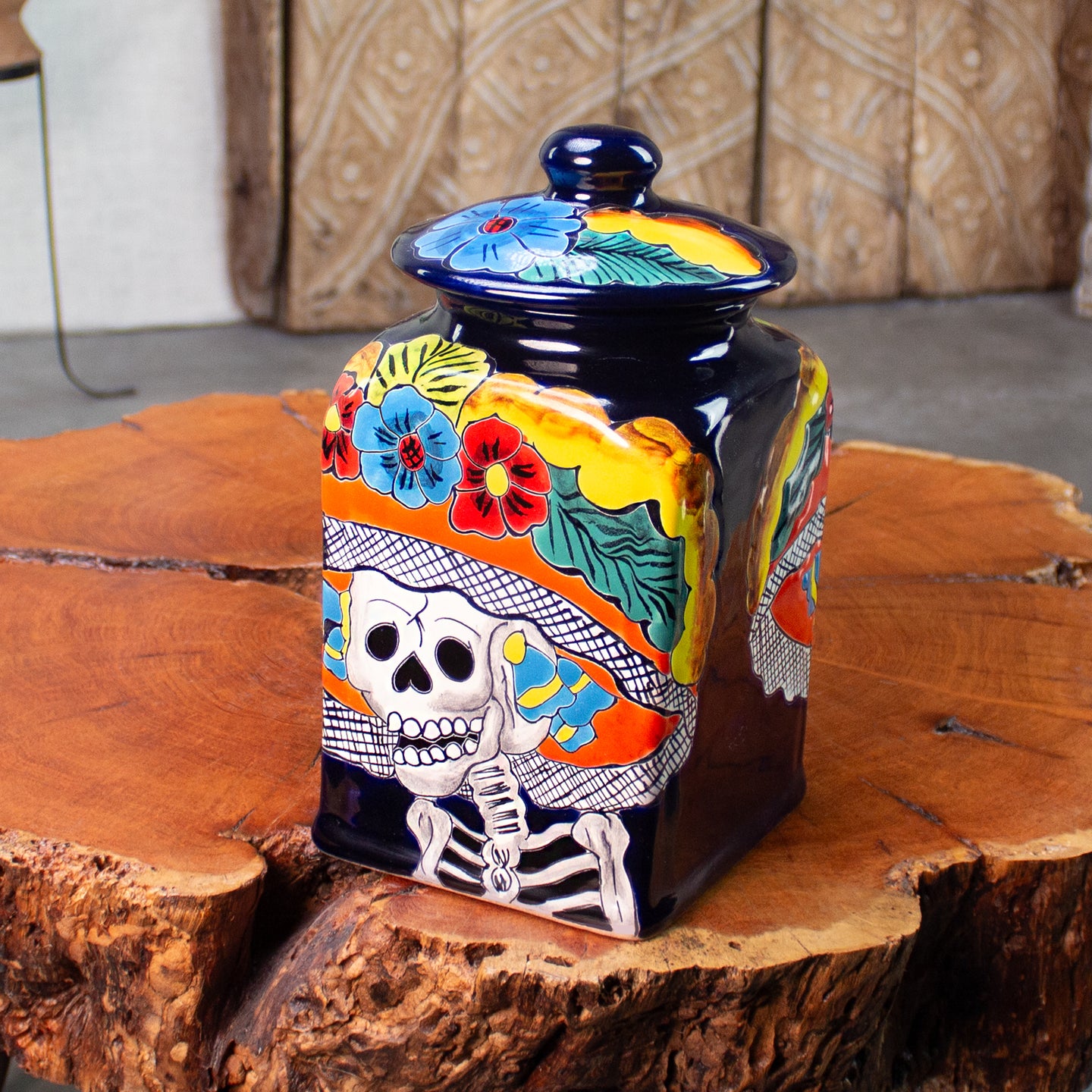 A hand-painted ceramic jar with a matching lid, decorated with a Day of the Dead theme featuring a skeleton wearing a floral hat. The canister is on a rustic wood table. 