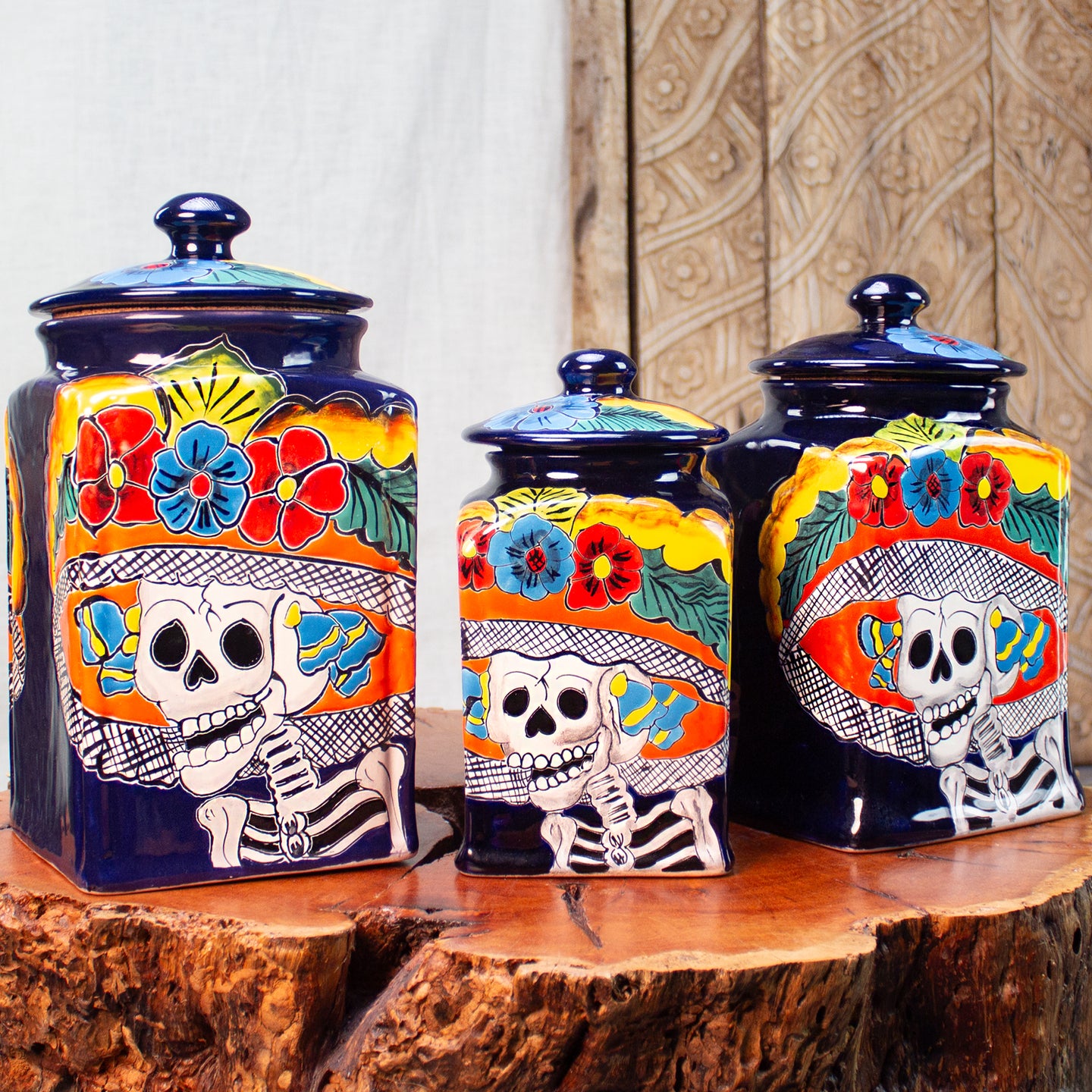 Three hand-painted ceramic canisters with matching lids, decorated with colorful floral and skeletal Day of the Dead motifs, displayed on a rustic wood table.