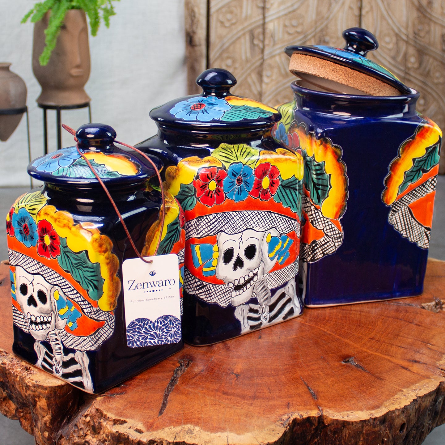 Three ceramic canisters hand-painted with Day of the Dead (Día de los Muertos) motifs, including sugar skulls and colorful flowers, displayed on a rustic wood table. A brand tag for 'Zenwaro' visible on the front canister.