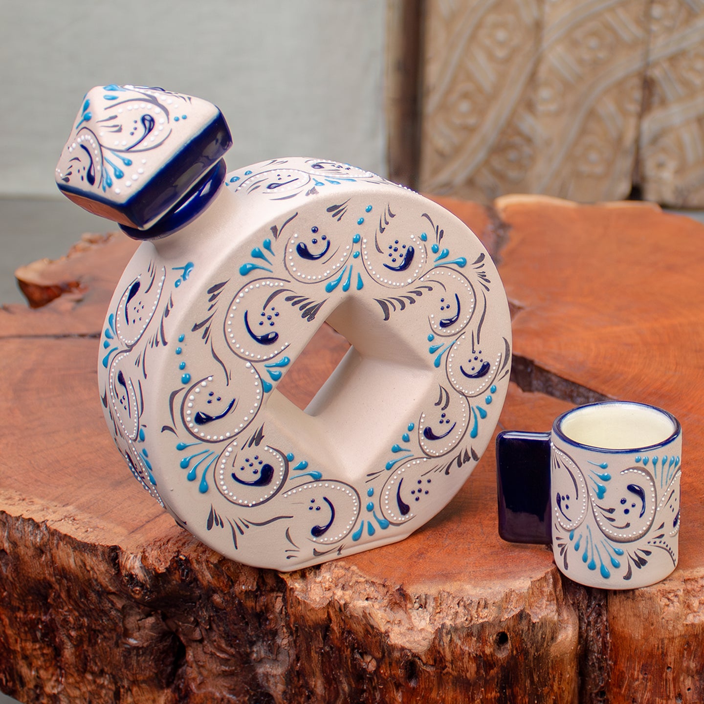 A hand-painted ceramic decanter with a matching small cup, decorated with blue and white floral patterns on an off-white base, displayed on a rustic wood table.