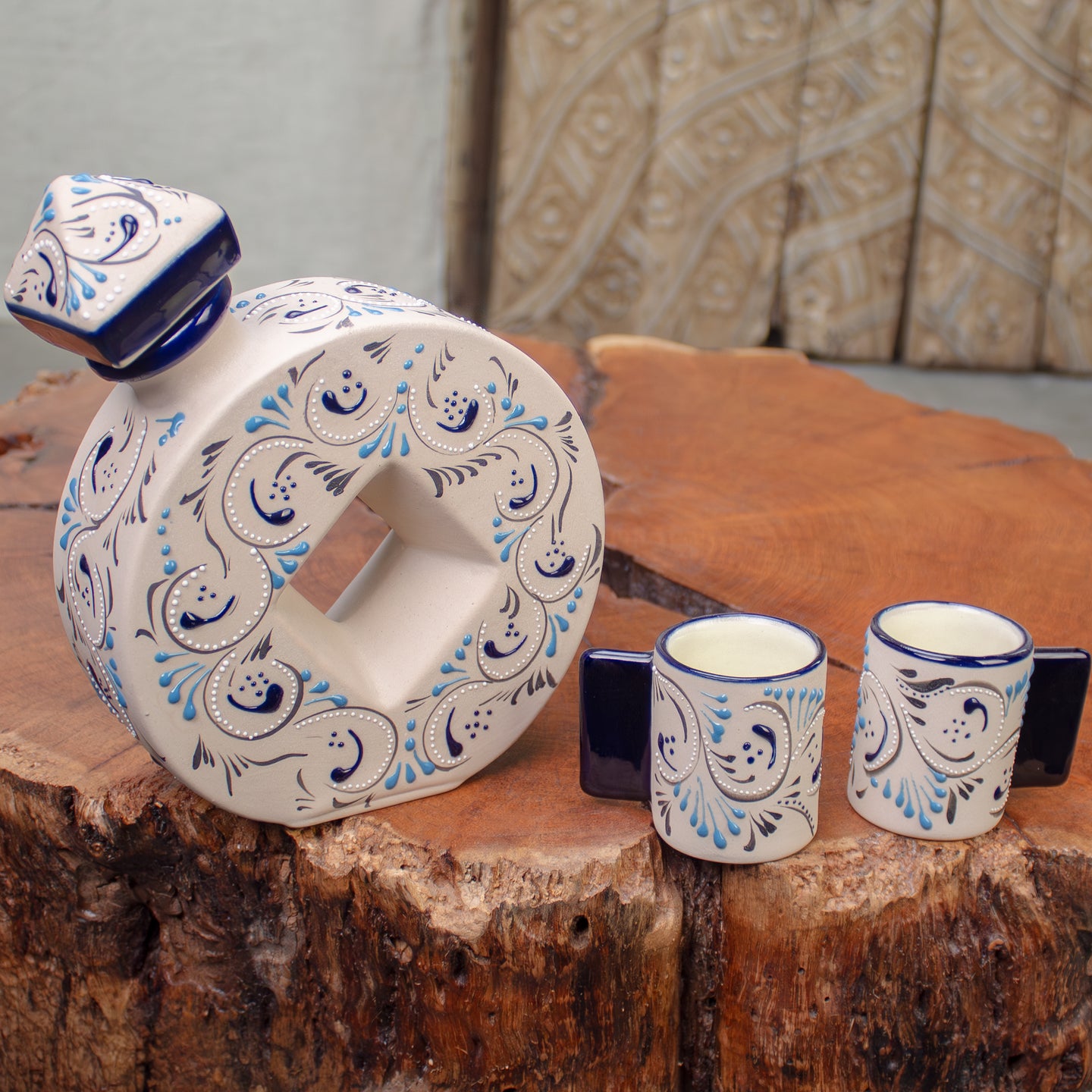A ceramic decanter with a matching small cup an, all decorated with a white base and blue and black floral scroll patterns, arranged on a rustic wood table.