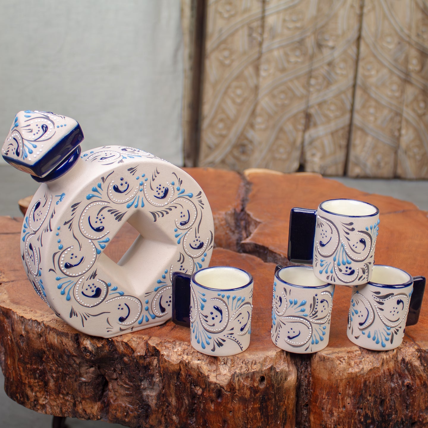 A ceramic decanter and four matching small cups with dark blue handles, decorated with a white base and blue and black floral scroll pattern, displayed on a rustic wood table.