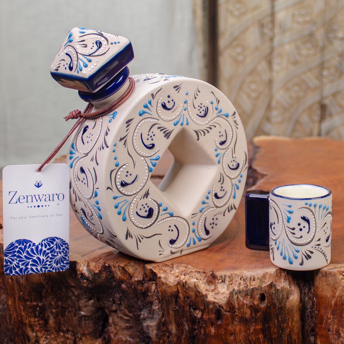 A decorative ceramic tequila bottle with a matching small cup and a Zenwaro branded hangtag, displayed on a rustic wood slab. The bottle and tumbler are cream-colored with blue and black paisley-style patterns, and the bottle is sealed with a blue ceramic stopper.