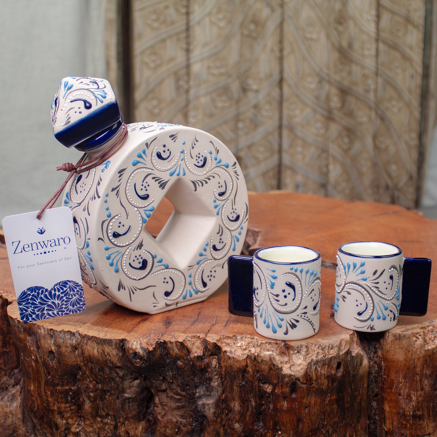 A ceramic liquor decanter and two matching small cups with a white base, blue rims, and blue paisley-style scrollwork, displayed on a rustic wood table with a Zenwaro branded hanging tag visible.