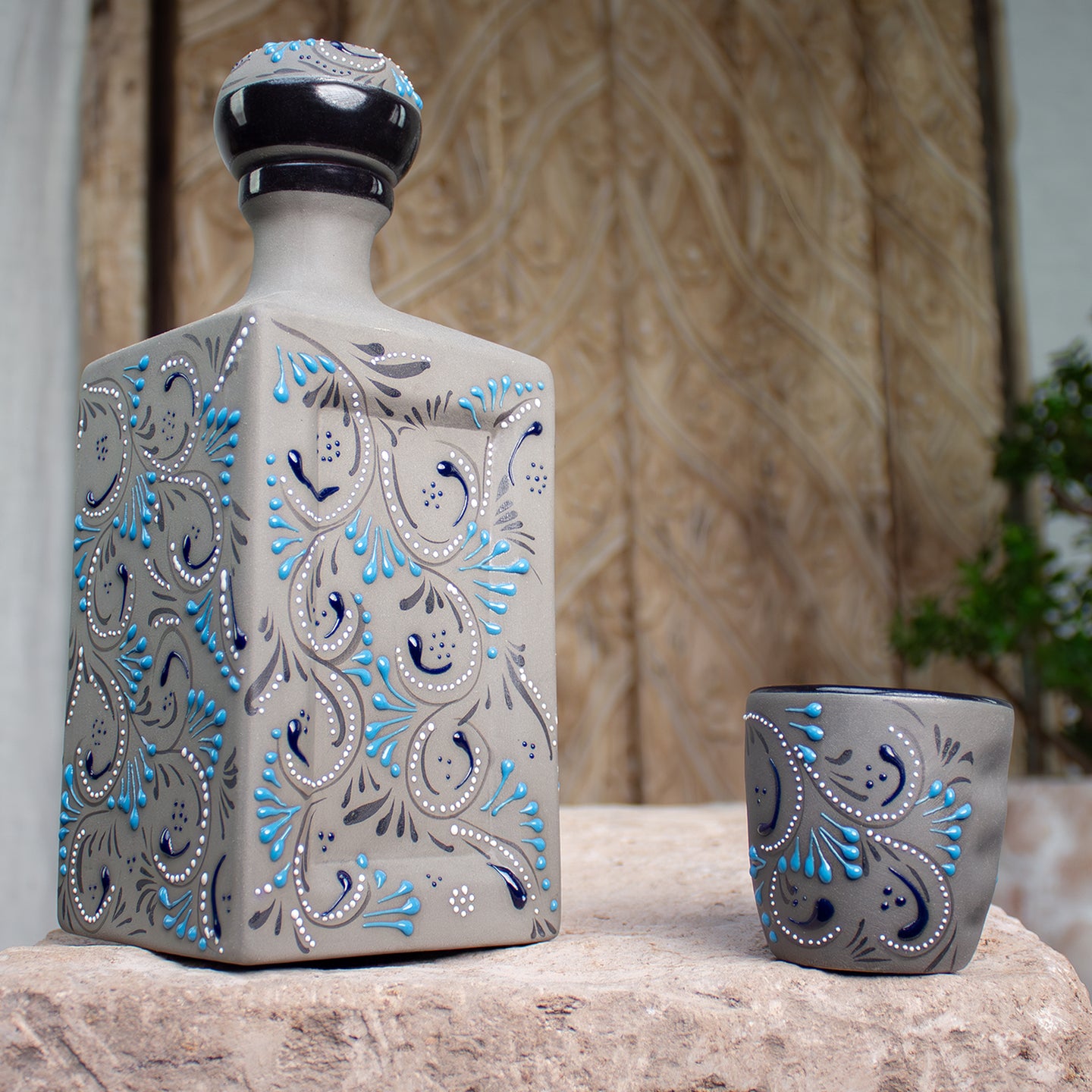 A decorative ceramic tequila bottle and matching shot glass set, featuring a gray base with hand-painted blue, black, and white paisley patterns, displayed on a stone counter.