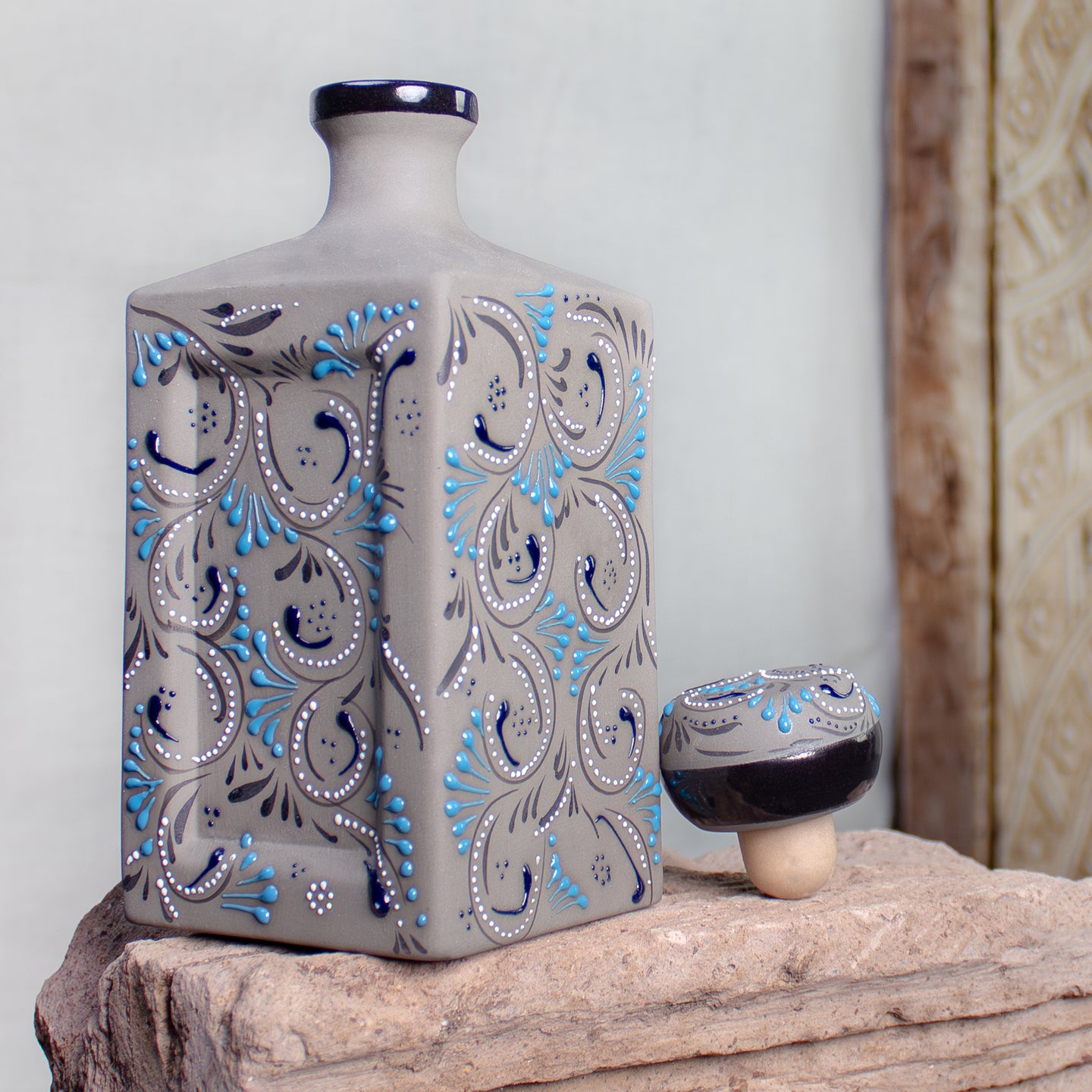 A square ceramic bottle with a narrow neck, decorated with a gray, black, and light blue paisley pattern, displayed on a rustic stone surface next to its matching cap.