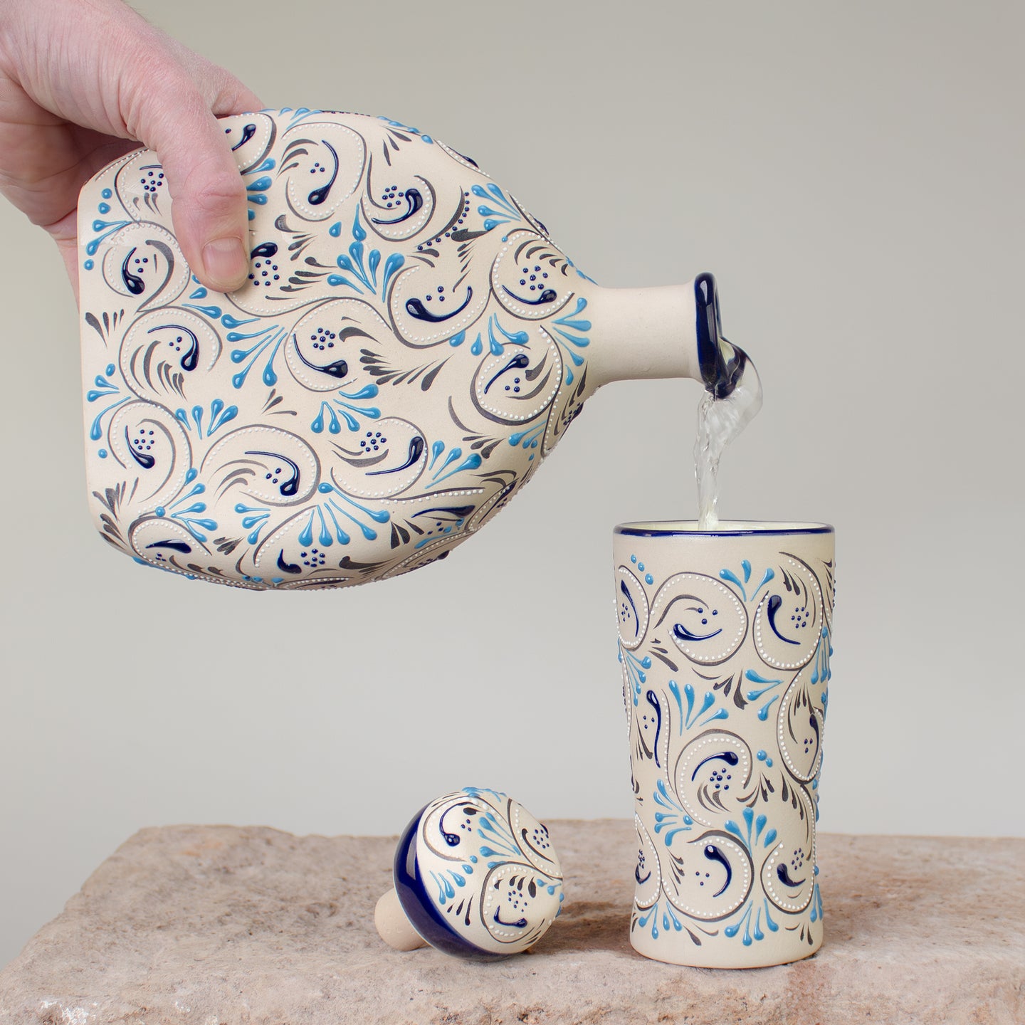 Ceramic decanter with decorative blue white  and black hand painted patterns being poured into a matching cup on a neutral background.