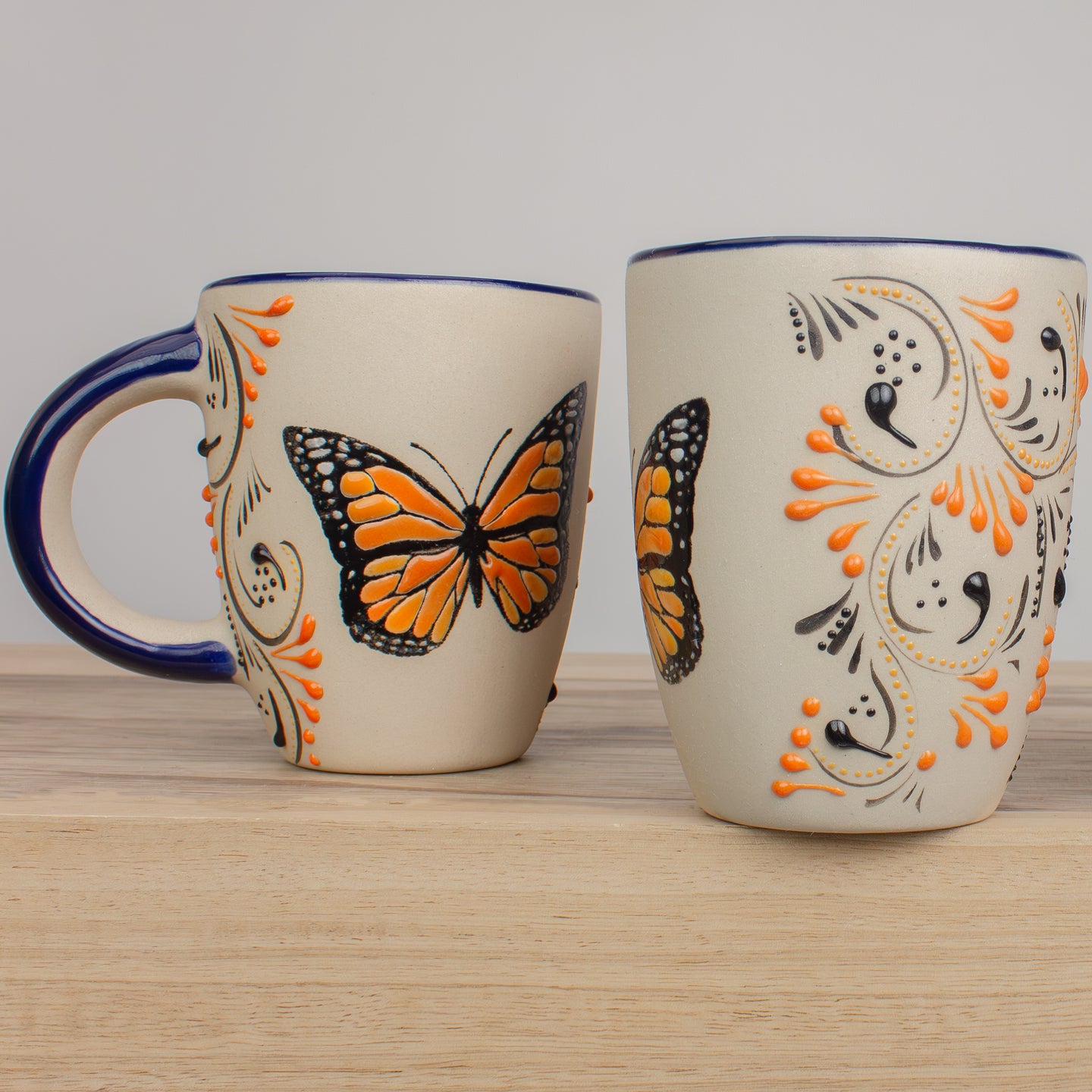 Two stoneware ceramic mugs with a vanilla-bean like color, hand painted with   raised orange and black  paisley patterns and an Monarch butterfly in center. A dark blue rim and handle, displayed on a light wood counter top.