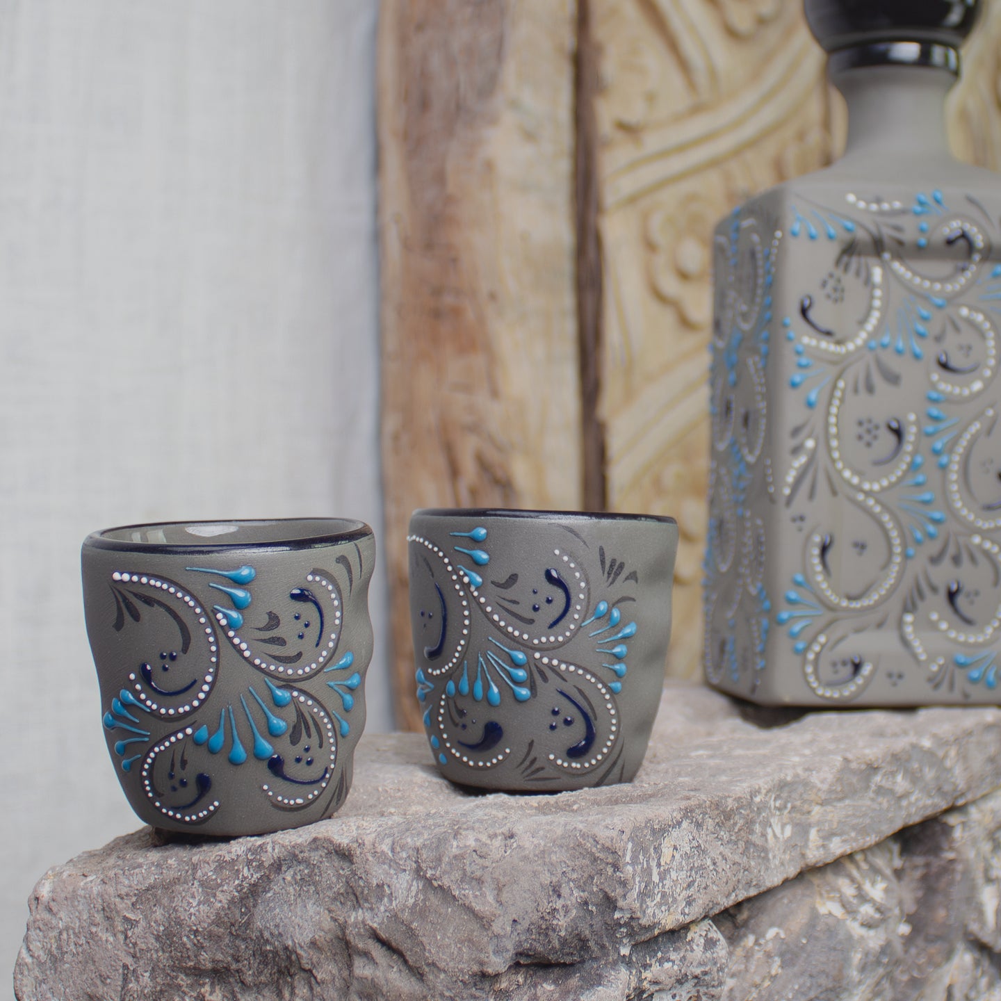 Two gray ceramic tumblers with blue and white paisley patterns sit on a stone surface next to a matching gray bottle with the same decorative pattern.