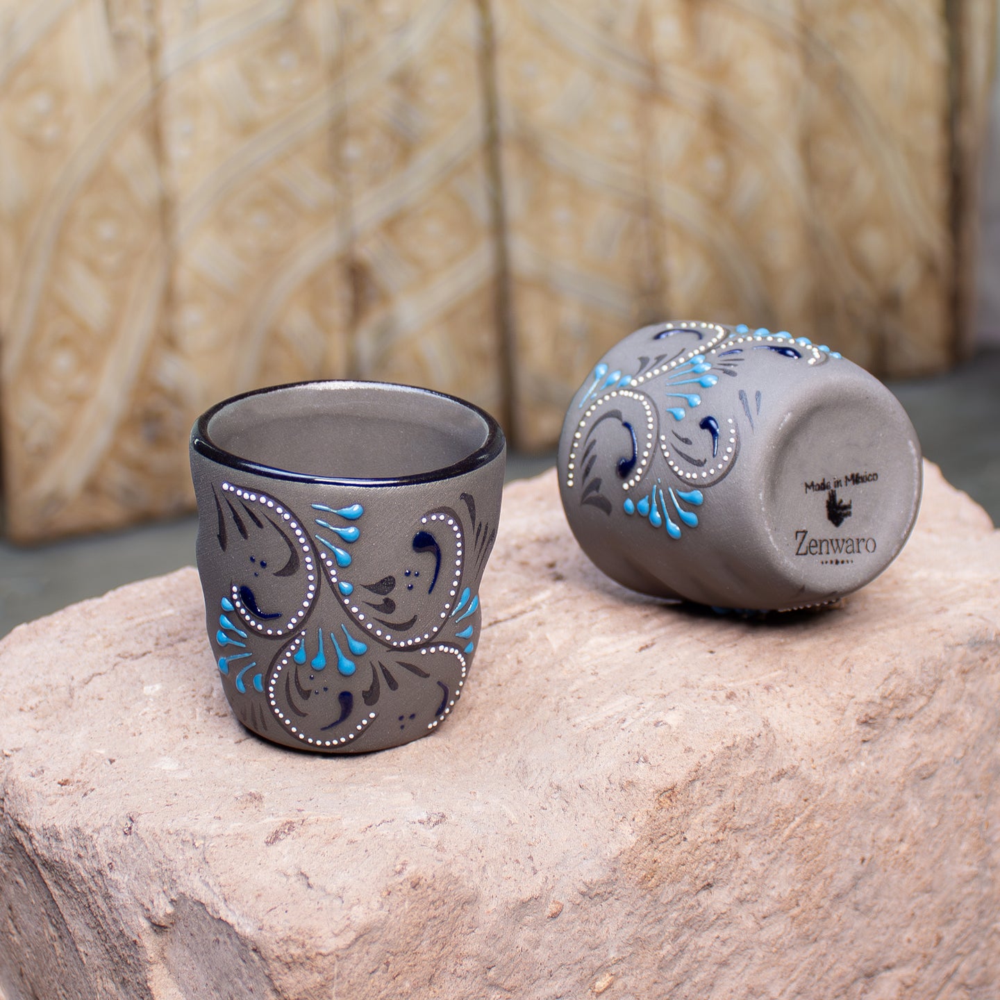 Two dark gray ceramic stoneware shot glasses with blue and white hand-painted floral designs, one standing upright and the other lying on its side showing the brand name Zenwaro, on stone counter.