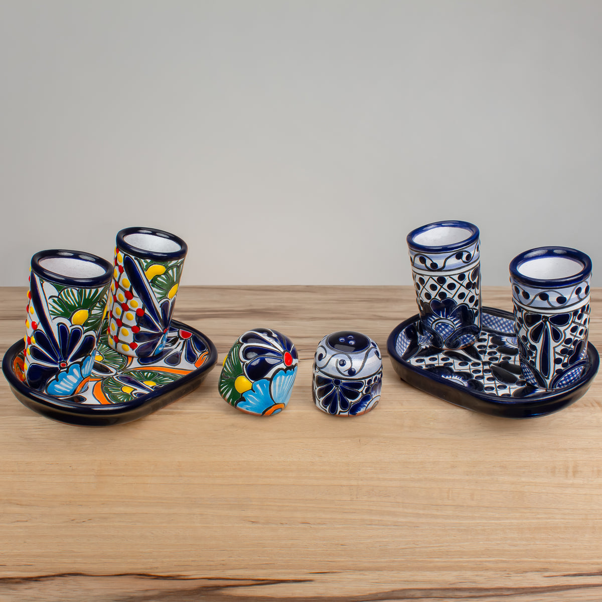 Two sets of hand-painted Talavera ceramic drinkware and barware displayed on a light wood surface against a neutral gray background. Each four piece set includes a shallow oval tray, two 4 ounce shot glasses, and a salt shaker, all decorated with a vibrant hand-painted motifs in blue, red, yellow, orange, and green on a white base.