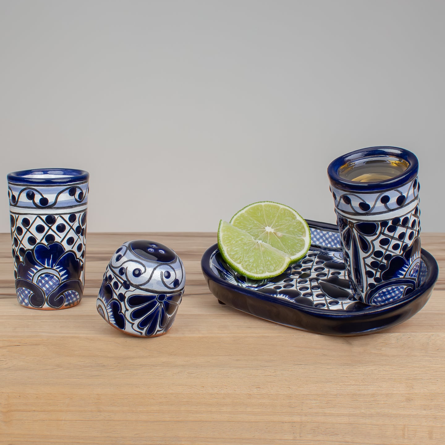 Three blue and white hand-painted ceramic tequila shot glasses with a traditional Mexican Talavera style, displayed on a light wood surface with a lime wedge on a tray.