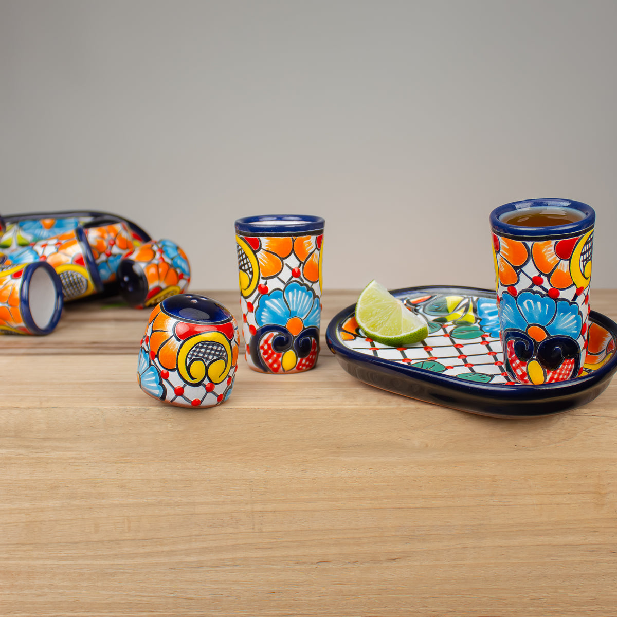 A set of hand-painted Mexican Talavera ceramic drinkware on a light wood table against a neutral gray background. The set includes a shallow oval tray, two 4 ounce shot glasses, and a salt shaker, all decorated with a vibrant floral and geometric pattern in orange, yellow, blue, red, and white with dark blue rims.