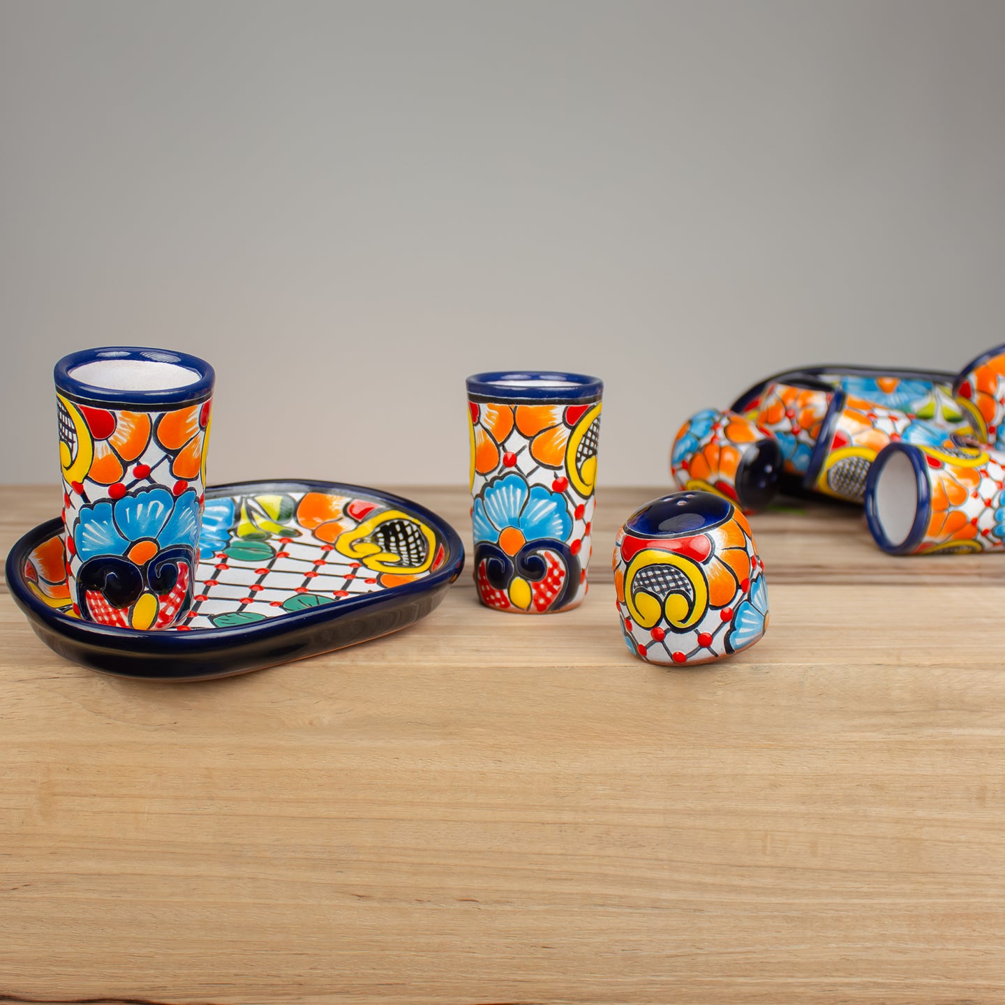 A set of hand-painted Mexican Talavera ceramic drinkware on a hard wood counter. Four piece set includes a shallow oval tray, two 4 ounce shot glasses, and a salt shaker, all decorated with a vibrant Talavera and geometric pattern in orange, yellow, blue, red, and white with dark blue rims.