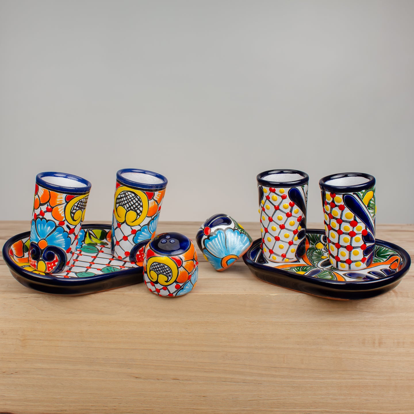 A colorful Mexican Talavera-style ceramic tableware set on a light wood surface against a neutral gray background. The set includes two oval trays, two patterned tumblers, a small bowl, a small jar, and a decorative shell-shaped object, all decorated with vibrant hand-painted motifs in blue, red, yellow, orange, and green on a white base.