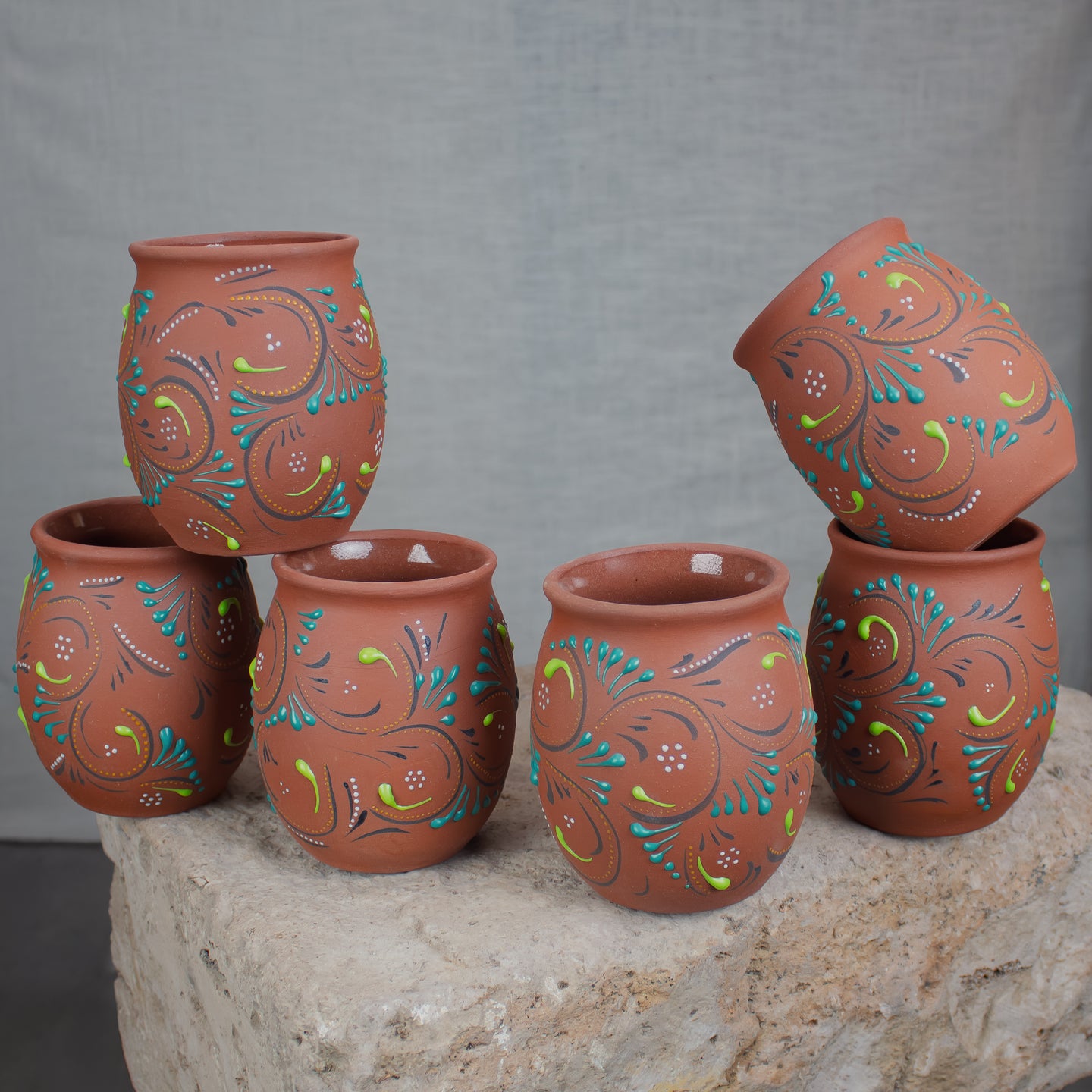 A set of six terracotta cocktail cups decorated with colorful hand-painted floral and scroll patterns, arranged on a stone table.
