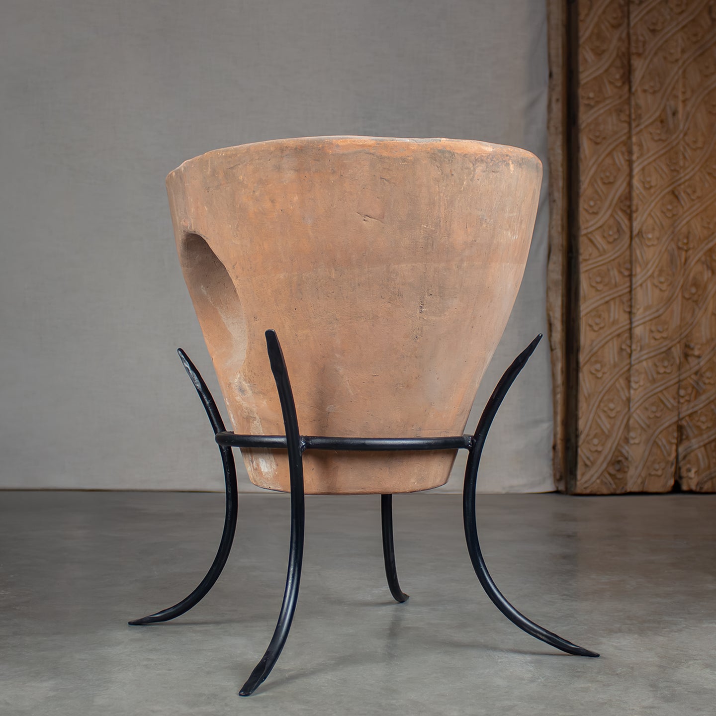 Large terracotta planter pot with carved out center, displayed on a black metal stand with curved legs, in front of carved wood door on concrete floor.