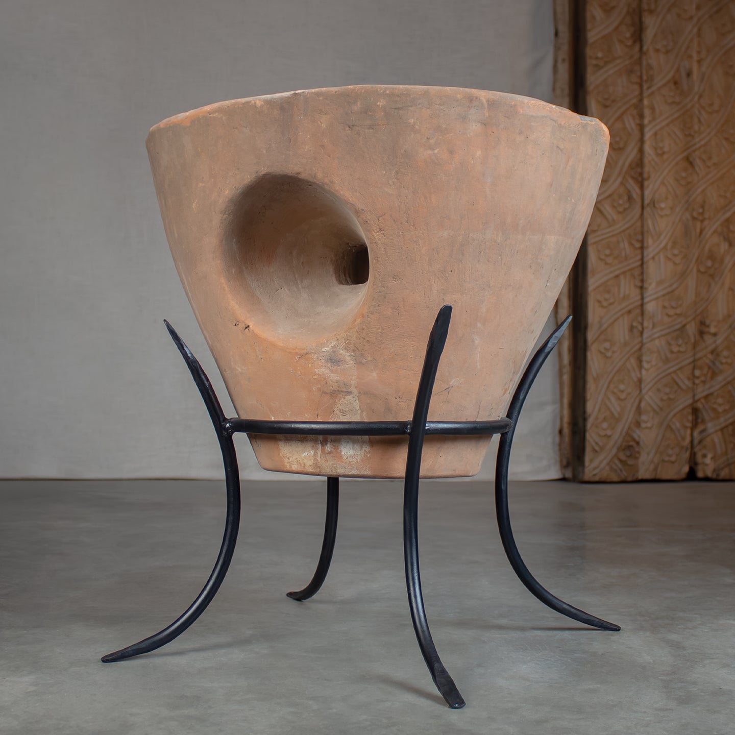 A terracotta clay planter pot with a large circular cutout, displayed on a black metal four-legged stand against a neutral gray studio background.