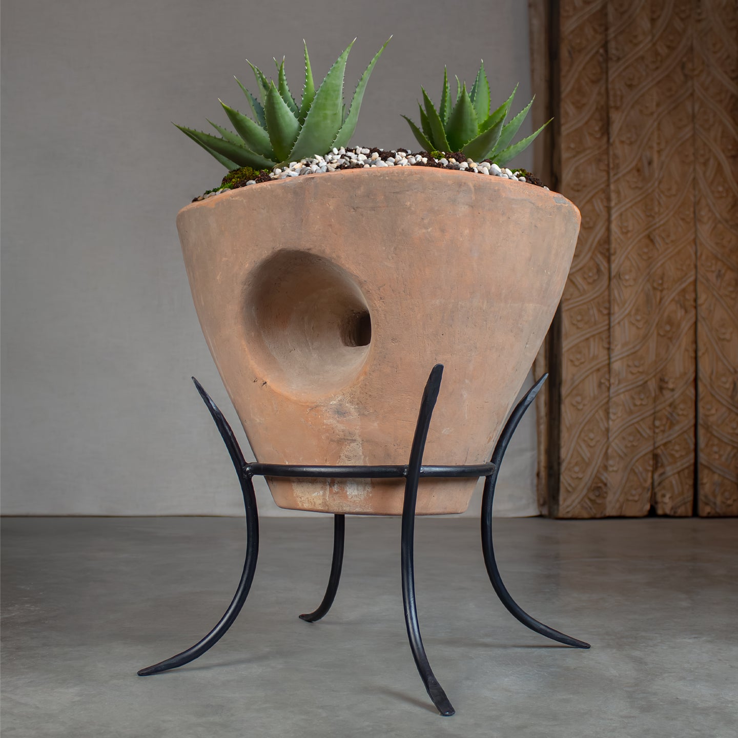 A large terracotta planter with a distinctive circular carveout through center, displayed on a black metal stand. The planter is filled with small white pebbles and two agave plants, in front of carved wood door on concrete floor.