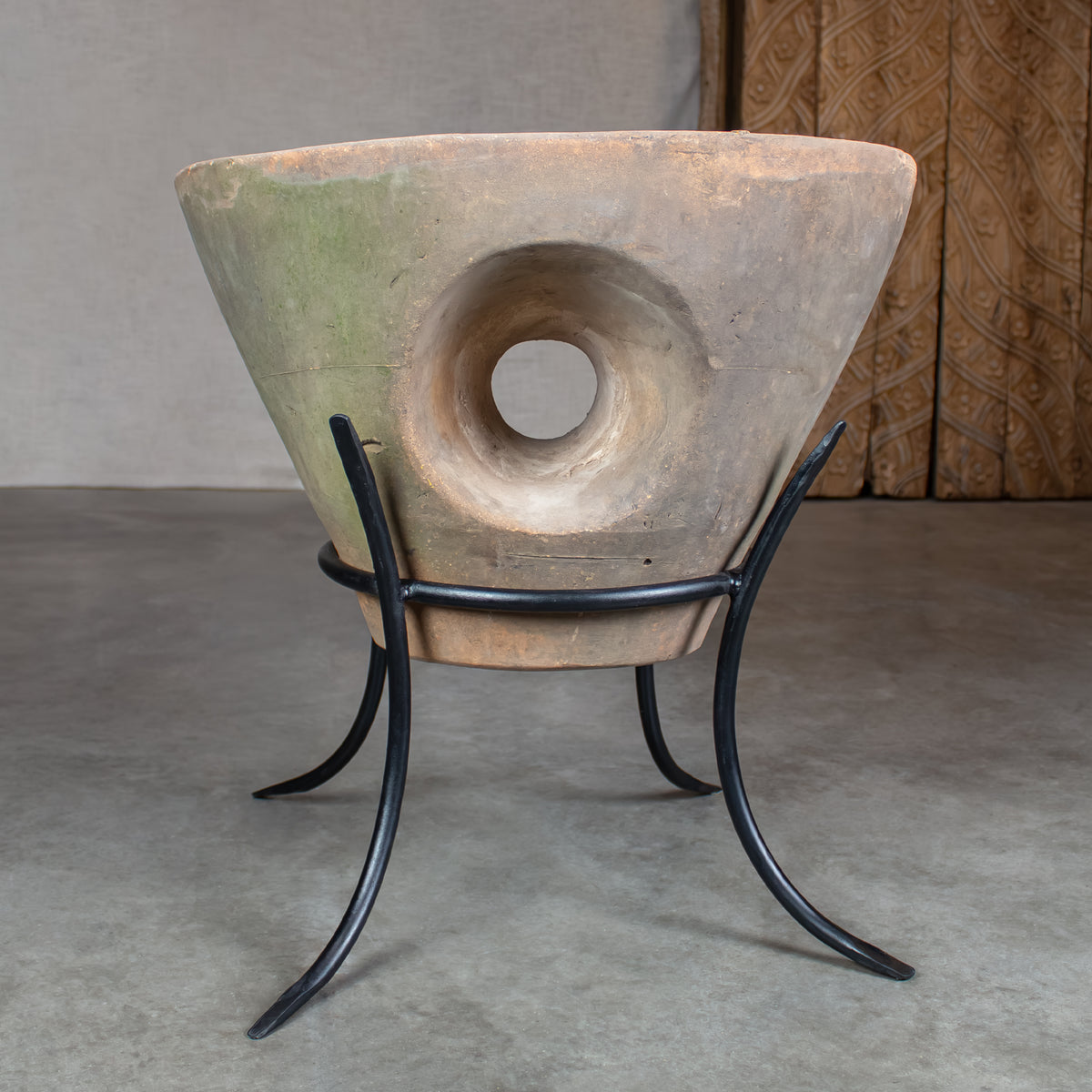 A large sculptural planter pot with a central circular cutout, displayed on a black metal stand. The planter has a textured, earthy coloration with greenish-gray on one side and a warm beige-brown on the other.