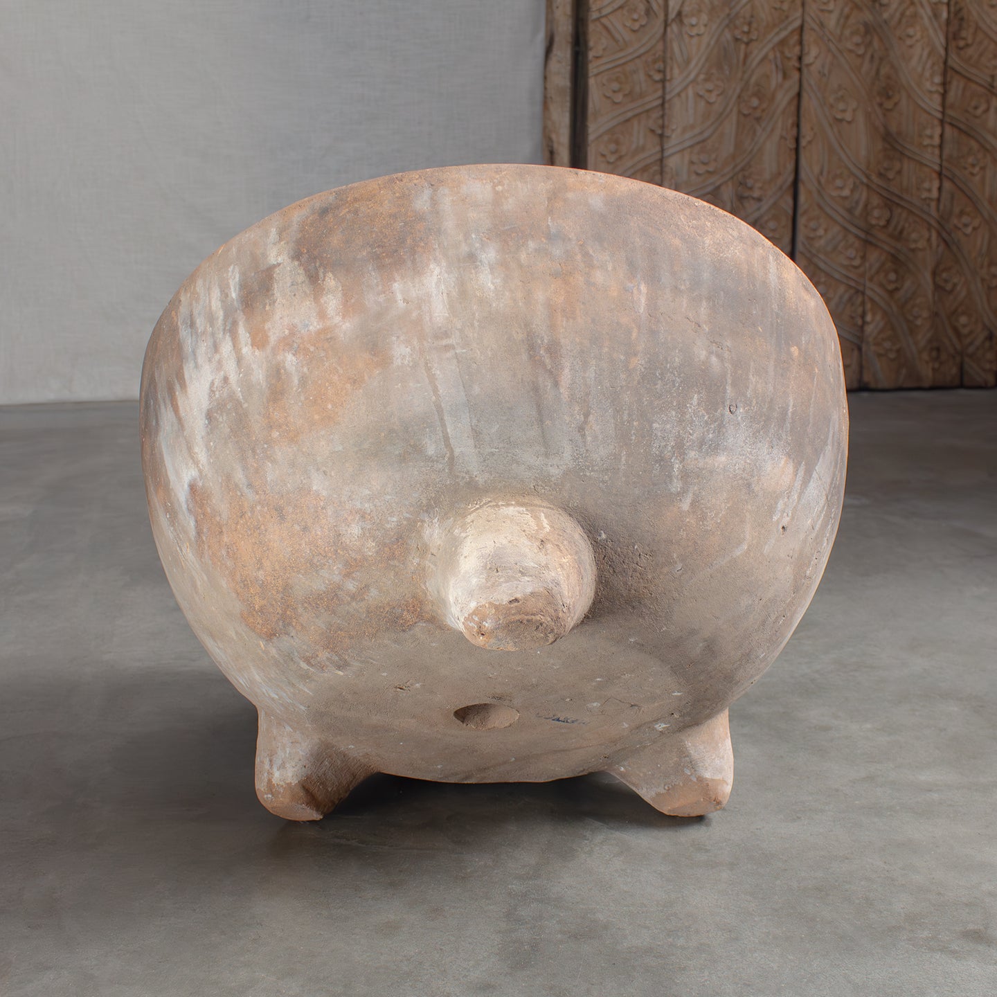 Vintage terracotta pot with feet on stained concrete floor in front of carved wood door.