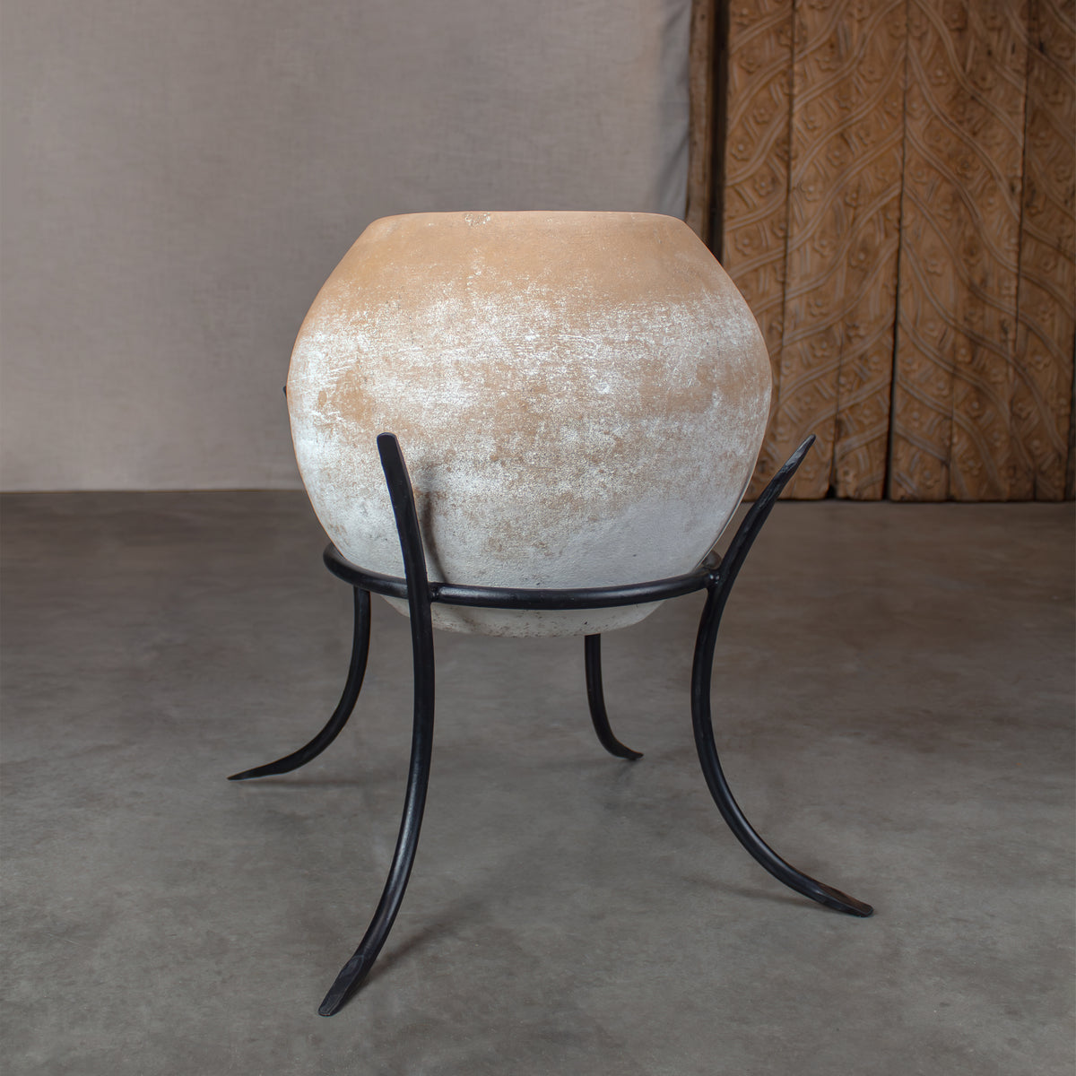 A large sphere terracotta planter pot with a distressed beige and white textured finish, displayed on a black metal stand with curved legs on concrete floor in front of carved wood door.