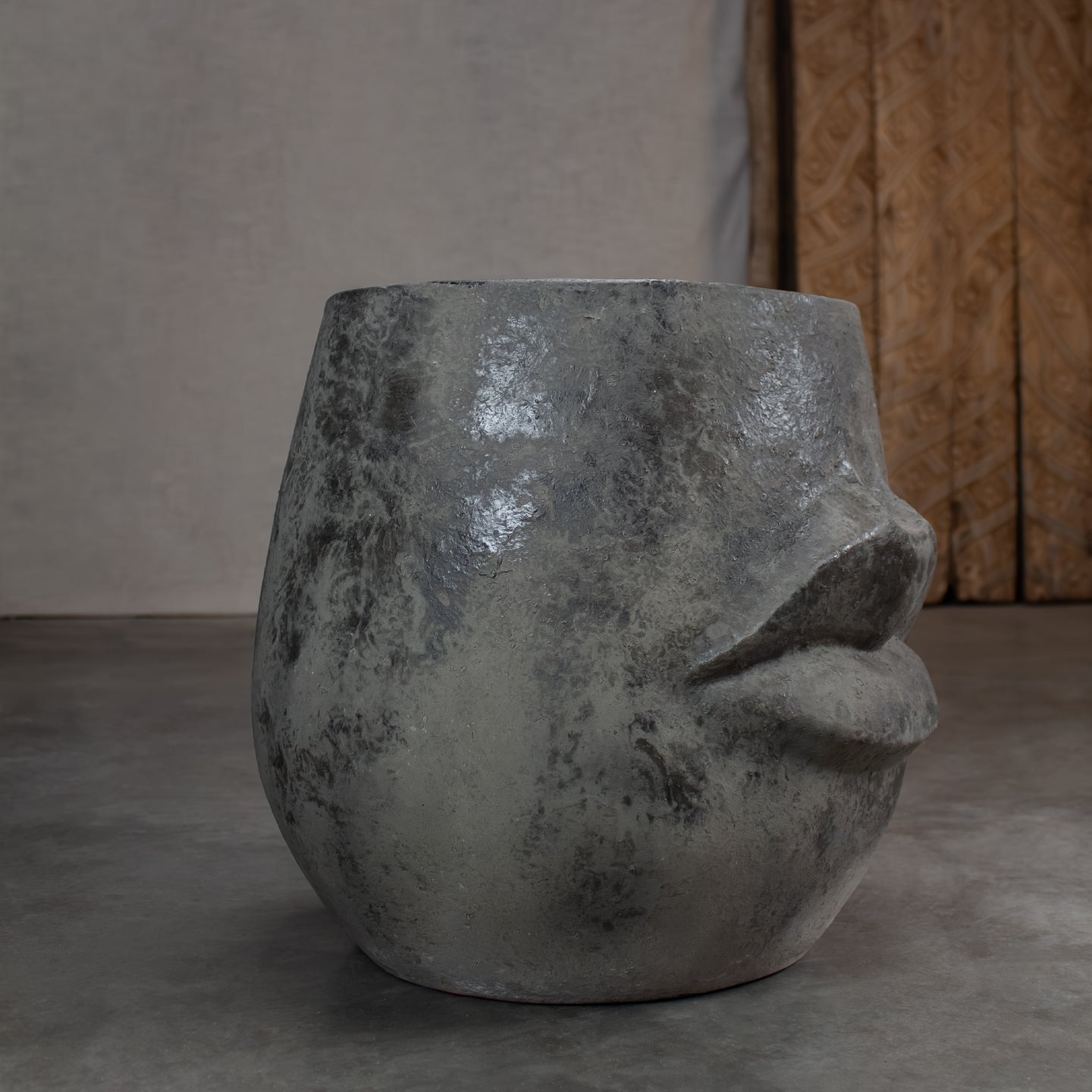 A sculptural terracotta pot with lips, shaped like a stylized face, on concrete patio in front of a carved wood door.