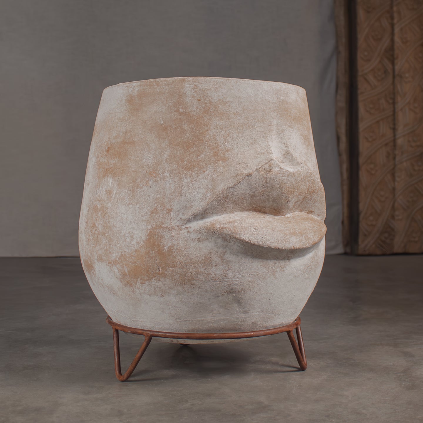 Abstract sculptural clay planter shaped like a stylized face, made of textured terracotta in beige and brown tones, sitting on a rustic iron pot stand with tripod hairpin legs in front of a carved wood barn door.