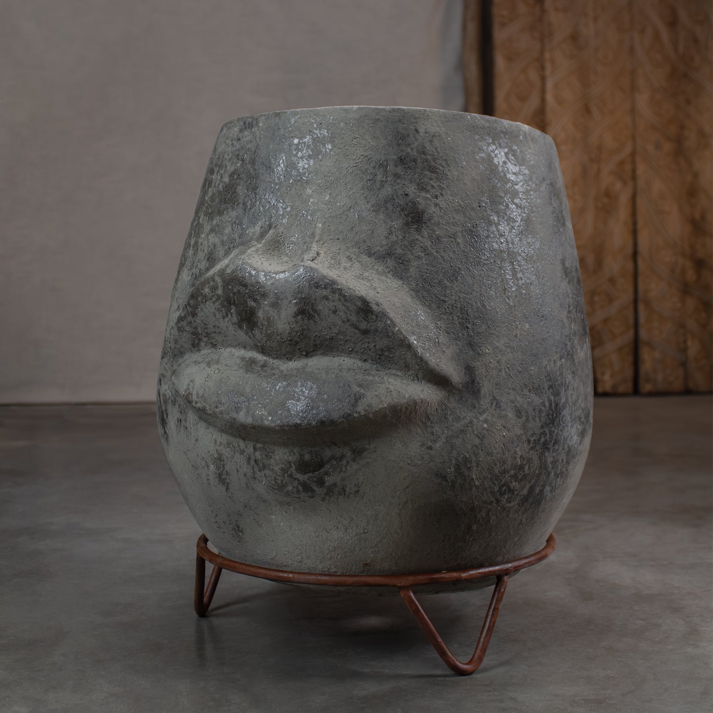 A sculptural black and gray planter shaped like a stylized face, resting on a metal stand against a carved wood door.
