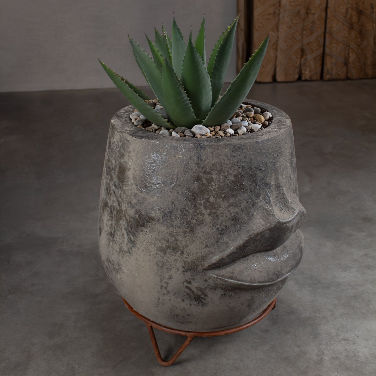 A green aloe vera plant in a textured gray concrete planter shaped like a face, sitting on a copper-colored metal stand against a gray concrete floor.
