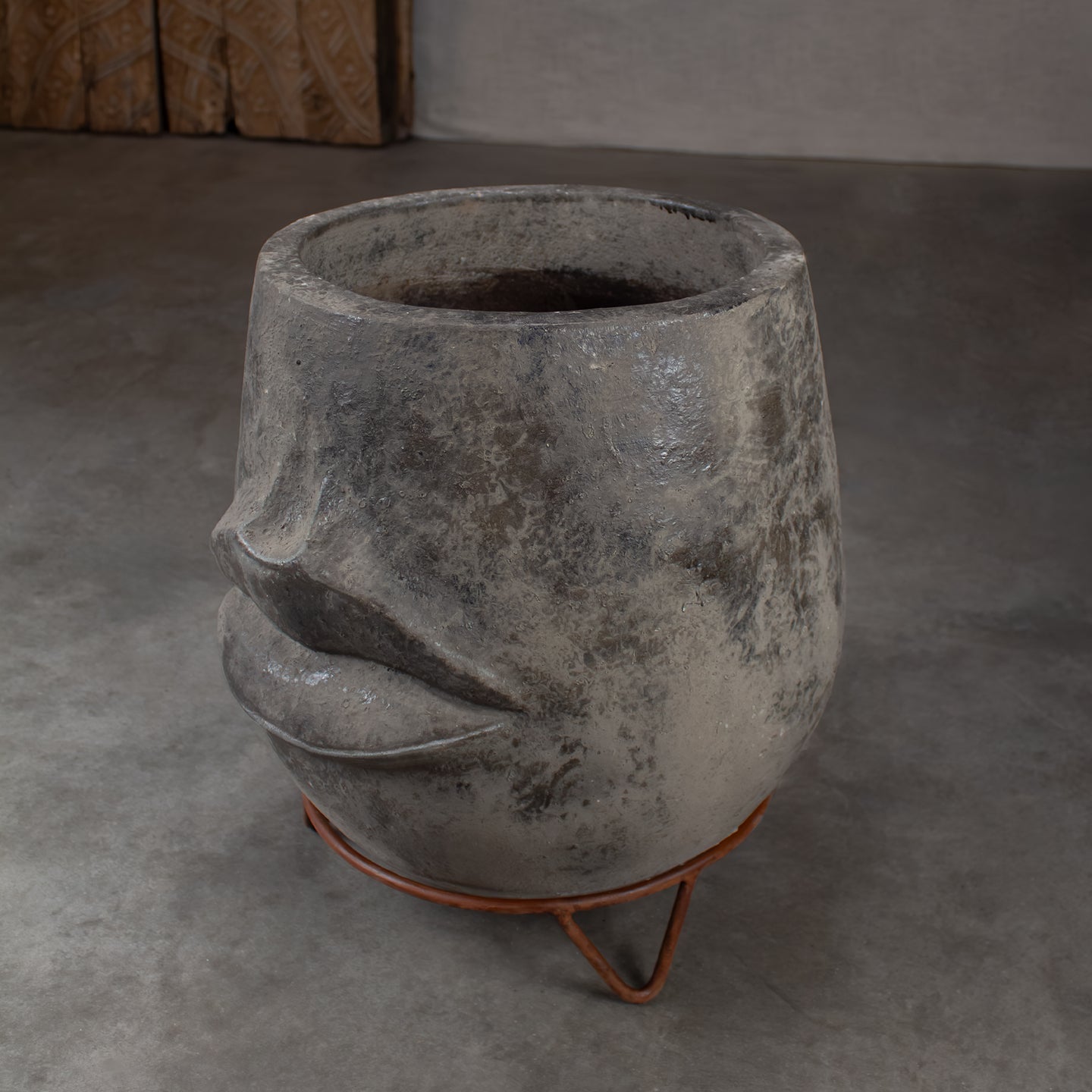 A sculptural terracotta planter shaped like a stylized face, on a rustic metal stand in a minimalist concrete setting.