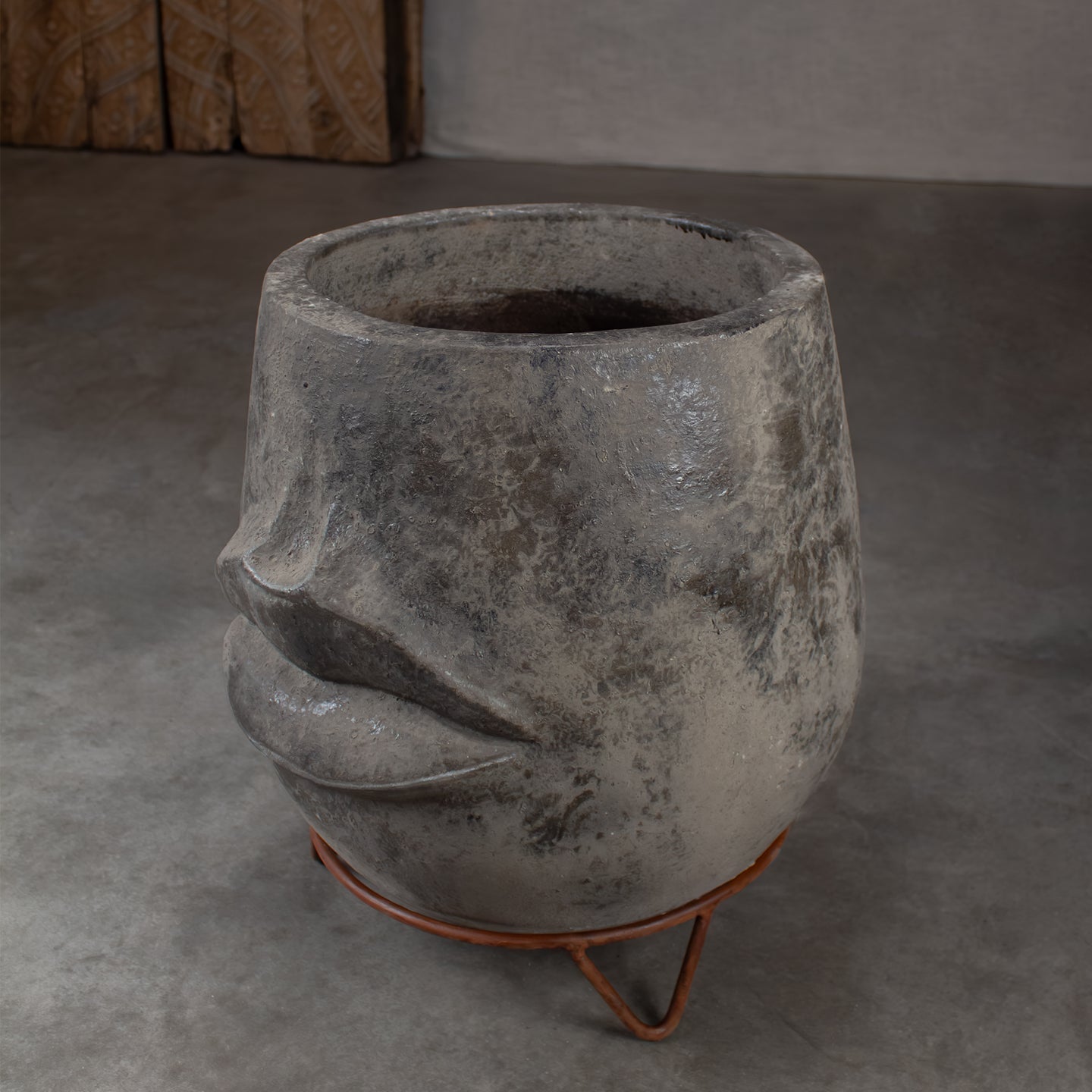A sculptural terracotta planter shaped like a stylized face, on a rustic metal stand in a minimalist concrete setting.