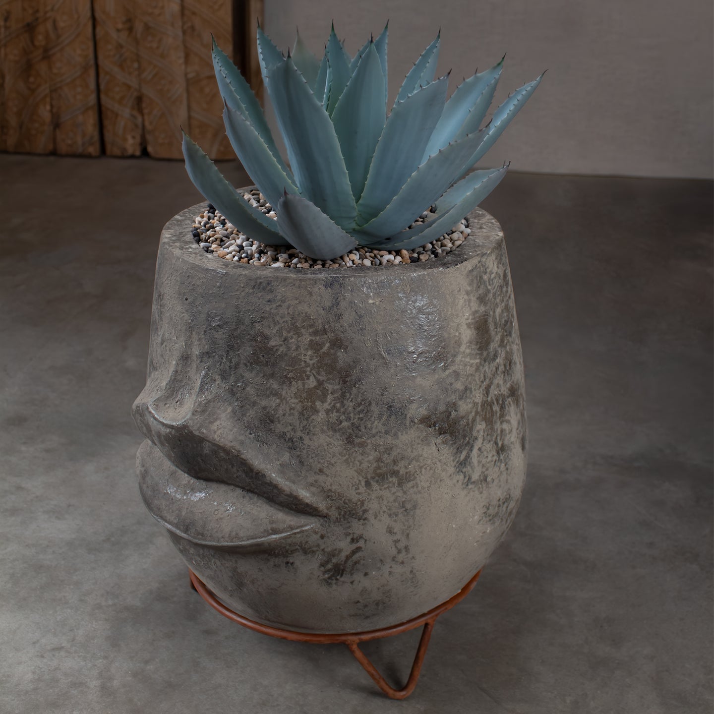 A blue-green agave succulent in a textured concrete planter shaped like a face, sitting on a iron stand on concrete floor.