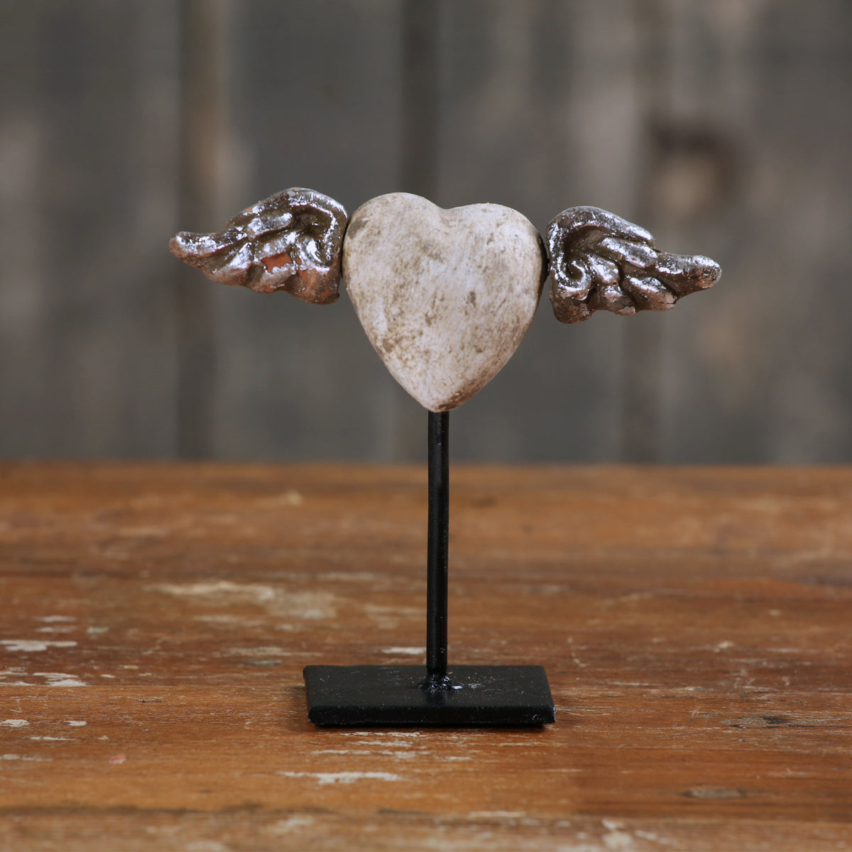 A small decorative heart sculpture with metallic angel wings on either side, mounted on a black stand on a rustic wooden surface.