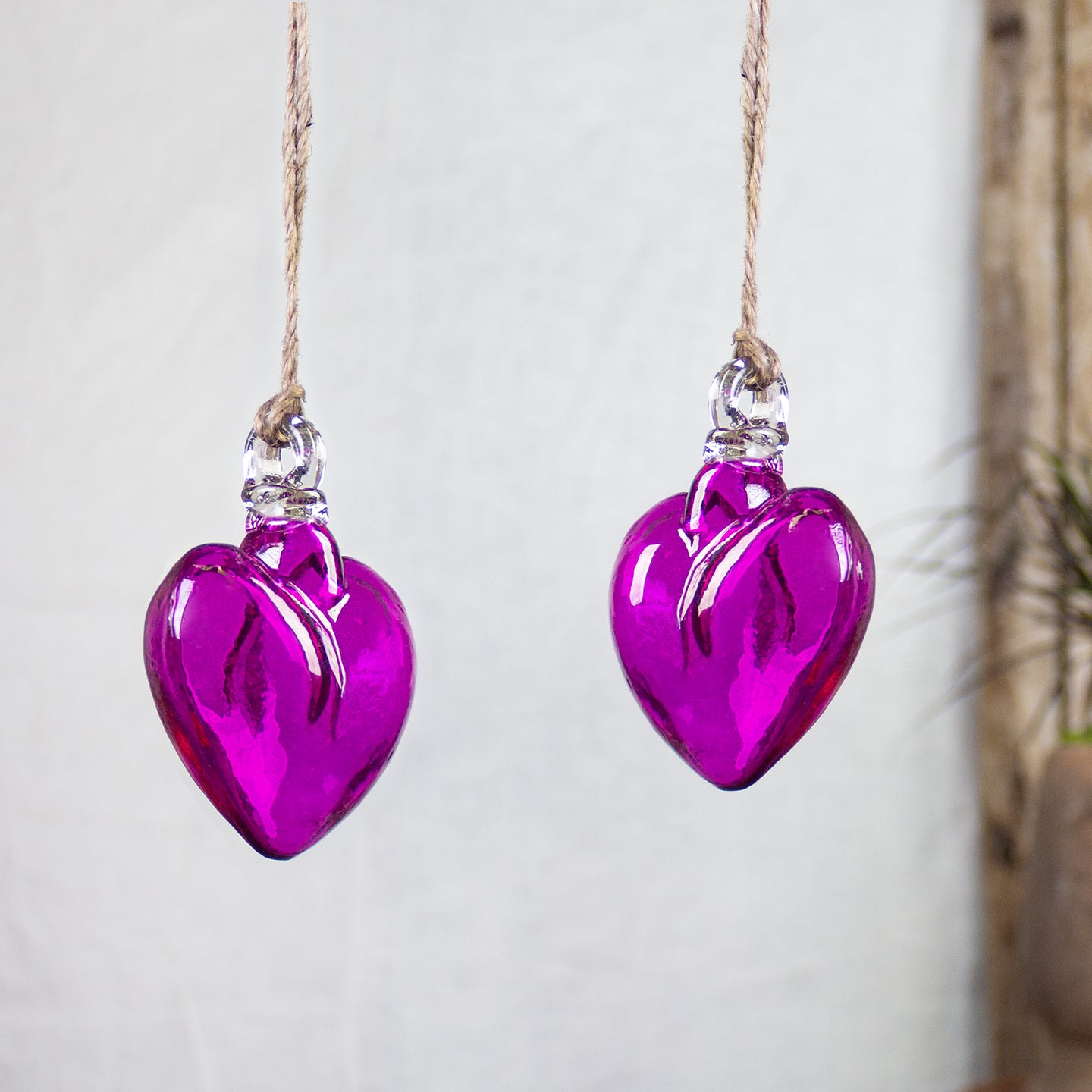 Two magenta heart-shaped glass ornaments hanging from natural jute twine against a soft, light gray textured background.