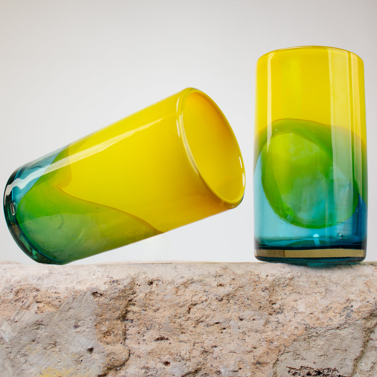 Two hand-blown glass tumblers with a vibrant yellow-to-green-to-blue gradient, displayed on a textured stone surface against a neutral light gray background.