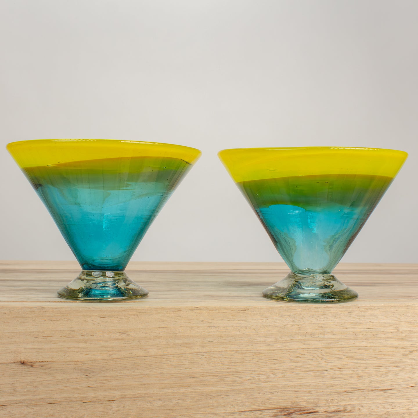 Two colorful Martini glasses with cosmic color transition from blue to green to yellow on a wooden counter. 