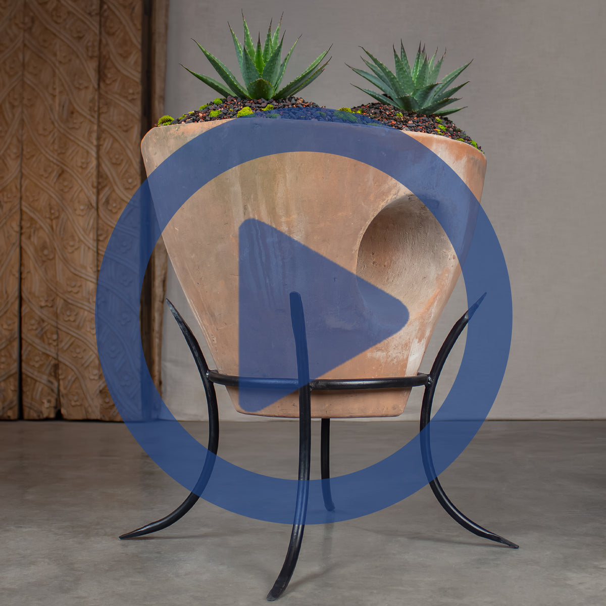Sculptural terracotta planter, displayed on a black metal pot stand. The planter is filled with dark gravel and two green Agave plants, in front of a carved wood.