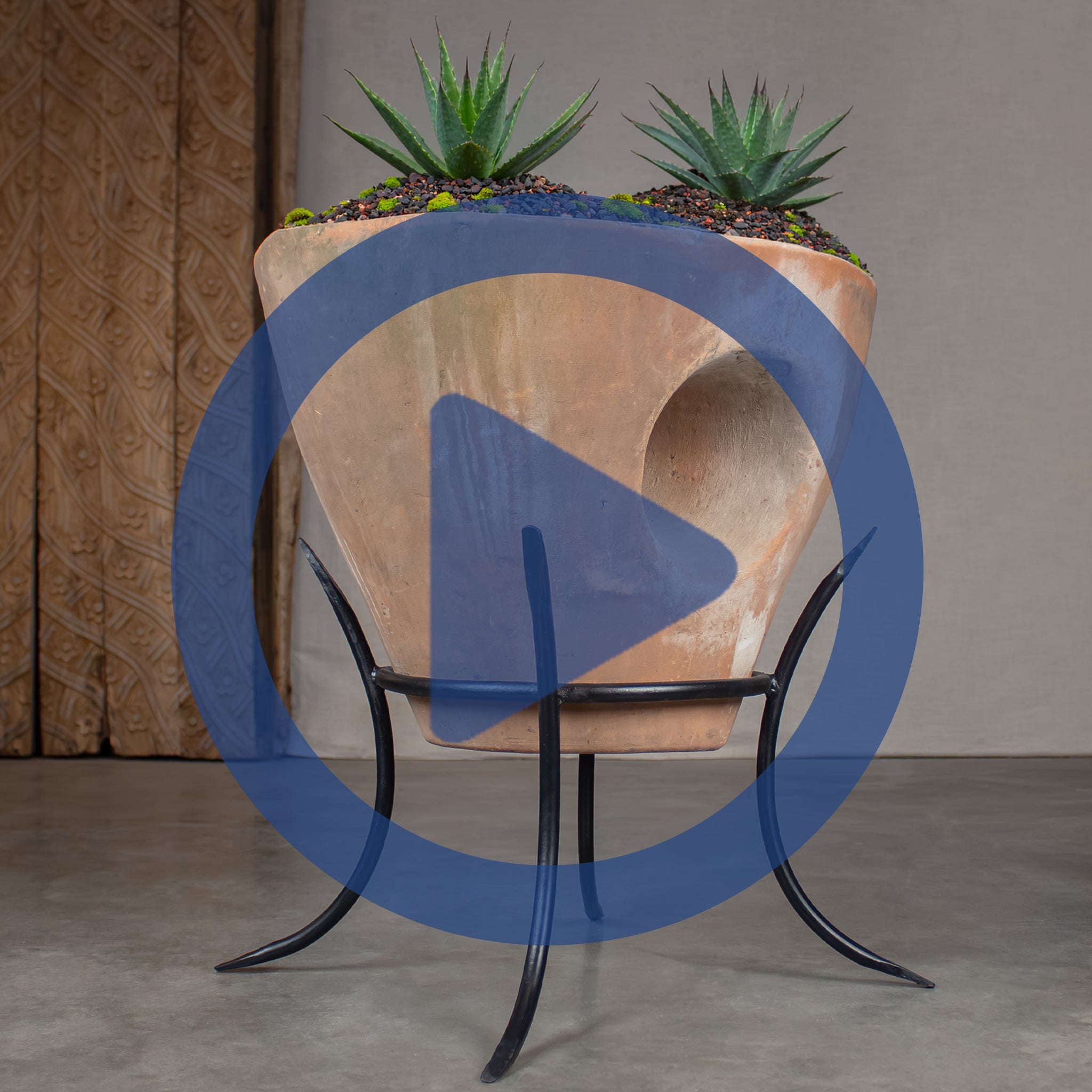 Sculptural terracotta planter, displayed on a black metal pot stand. The planter is filled with dark gravel and two green Agave plants, in front of a carved wood.